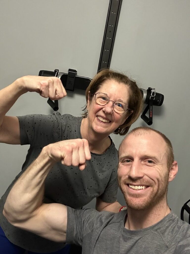 Two people smiling and flexing their arm muscles in a gym.